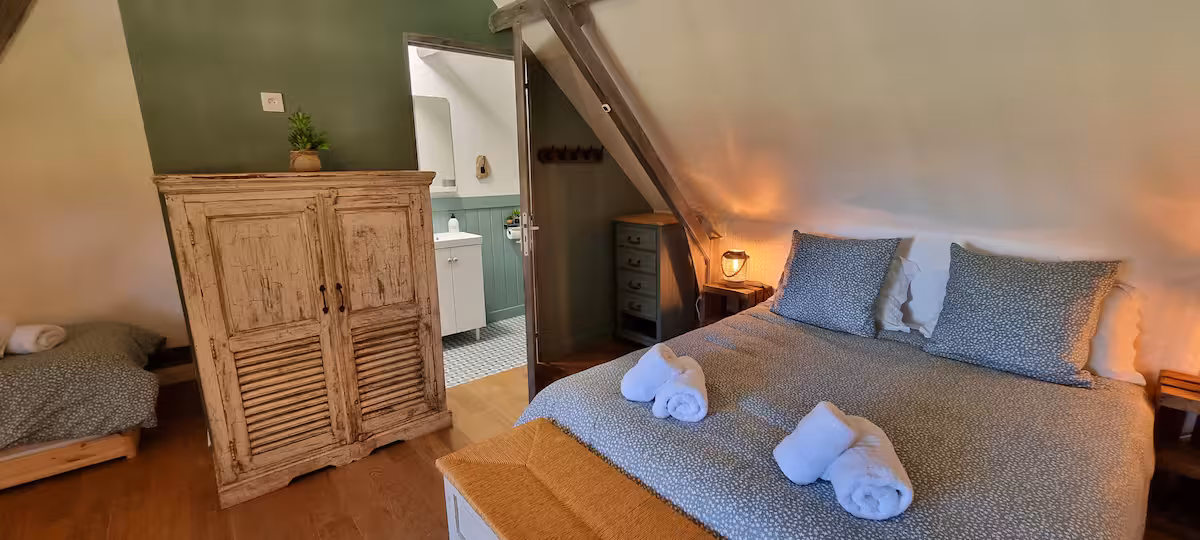 Chambre 1 sous combles : un grand lit avec linge fleuri bleu, commode patinée et accès à la salle de bain attenante.