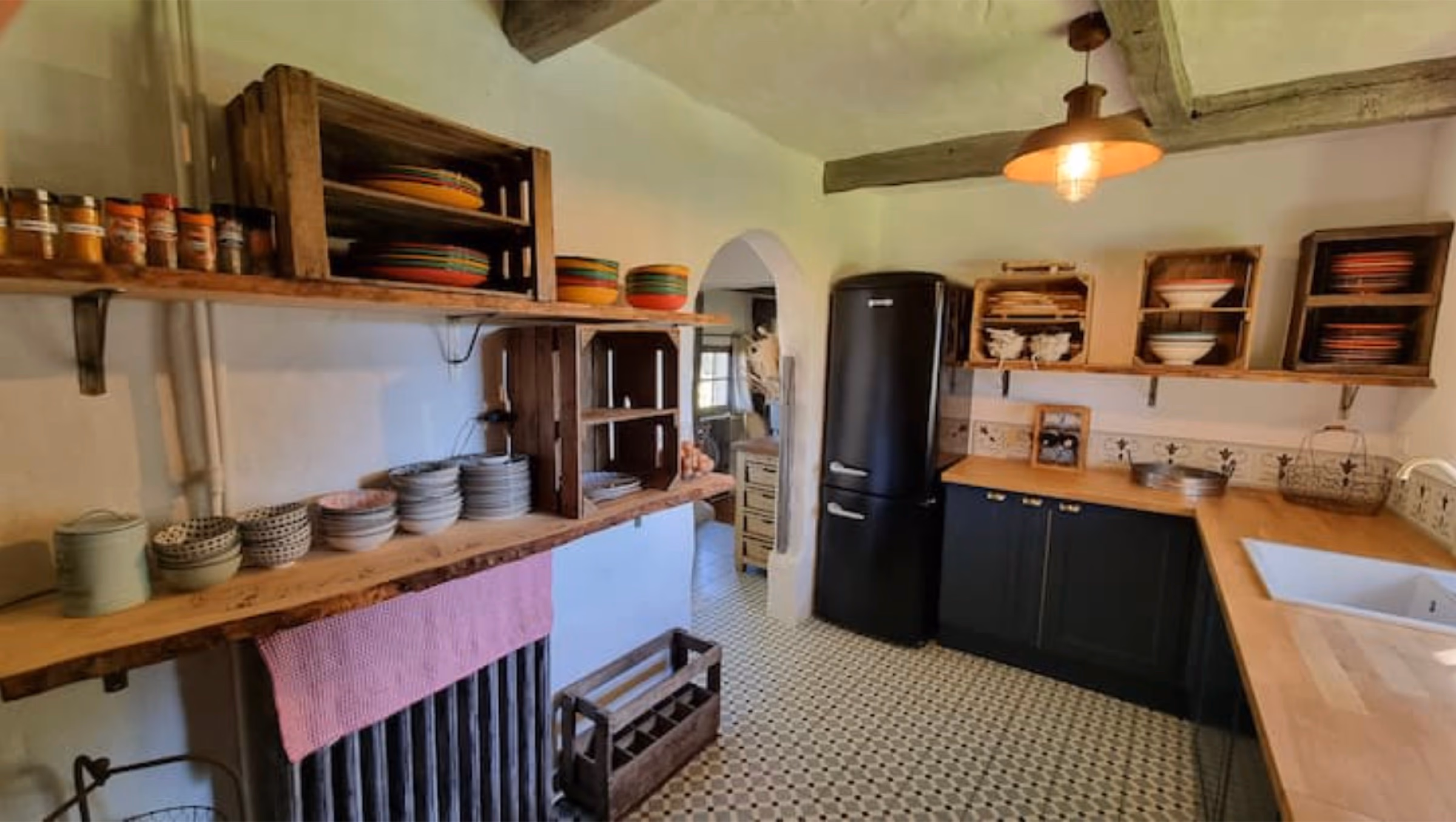 Cuisine équipée à la décoration normande : étagères ouvertes en caisses de bois, vaisselle colorée, frigo rétro noir et sol en carreaux de ciment damier.