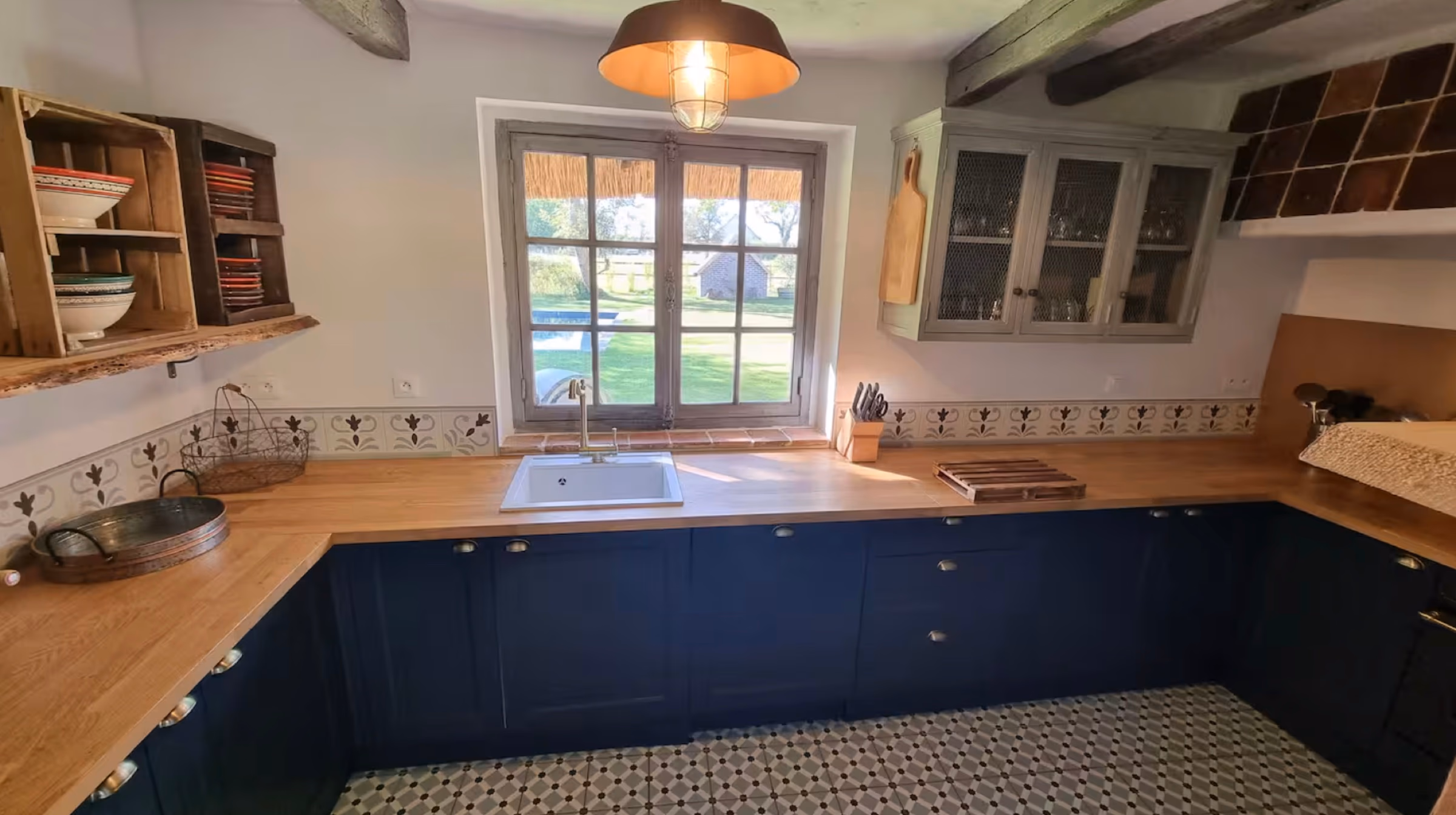 Plan de travail en bois massif sur meubles bleu nuit, lavabo, fenêtre à petits carreaux donnant sur le jardin et la piscine.