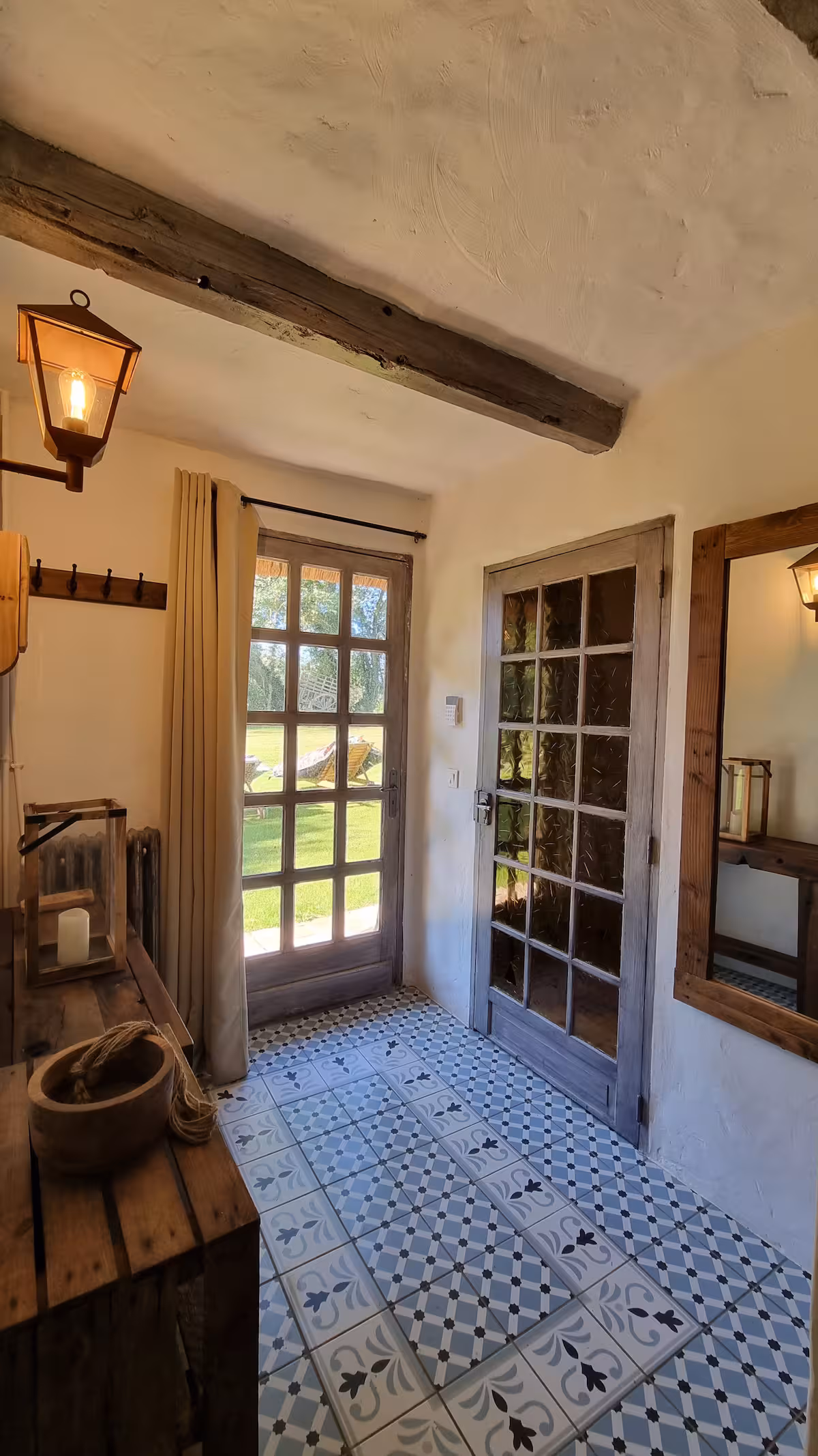 Entrée avec porte vitrée donnant sur le jardin, sol en carreaux de ciment, lampe lanterne en applique et poutre patinée au plafond.