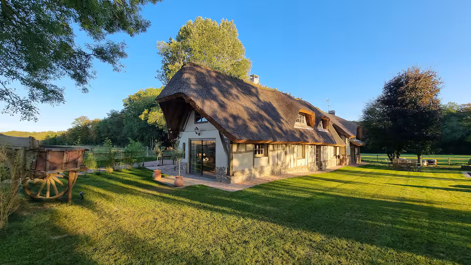 Façade de la chaumière normande au toit de chaume avec colombages, vue trois-quarts au coucher du soleil. Une ancienne charrette en bois complète le décor du jardin.