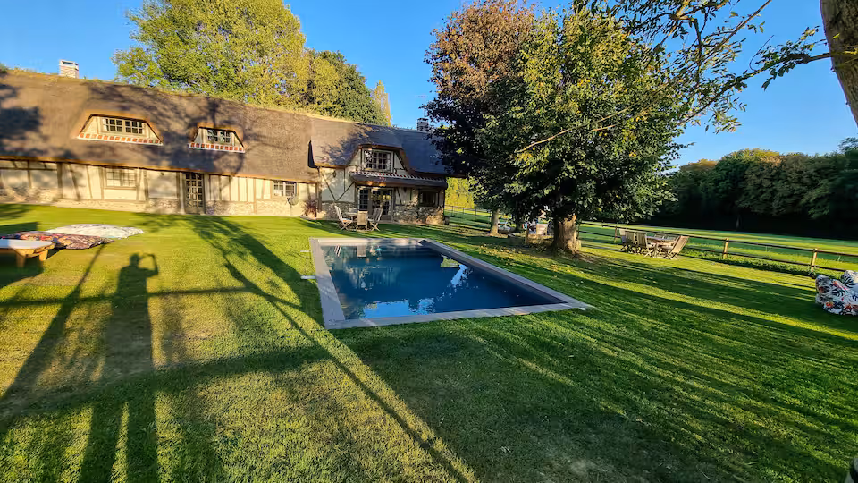Façade de la chaumière au toit de chaume reflétée dans la piscine, ombres longues du soir.