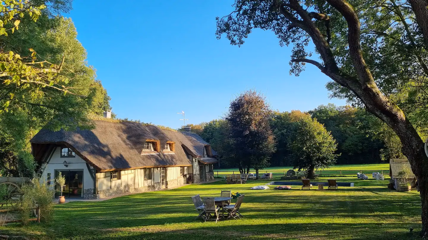 Vue grand-angle du parc avec la chaumière au fond, le jardin tondu, une grande table conviviale et la piscine au loin sous un ciel bleu.