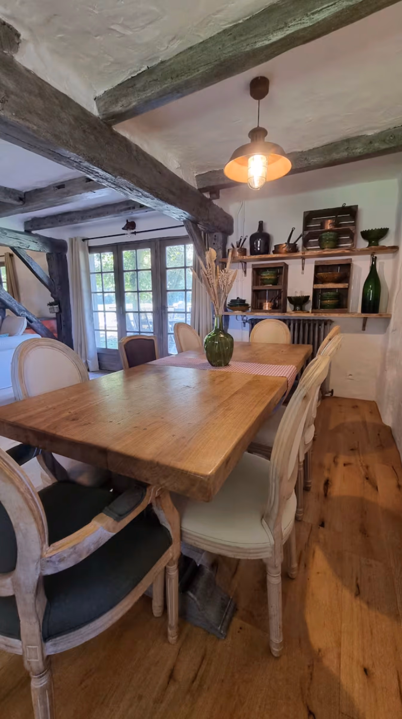 Grande table en bois rustique avec chaises médaillon, lampe industrielle suspendue, étagères avec poteries vertes anciennes et porte-fenêtre vers le jardin.
