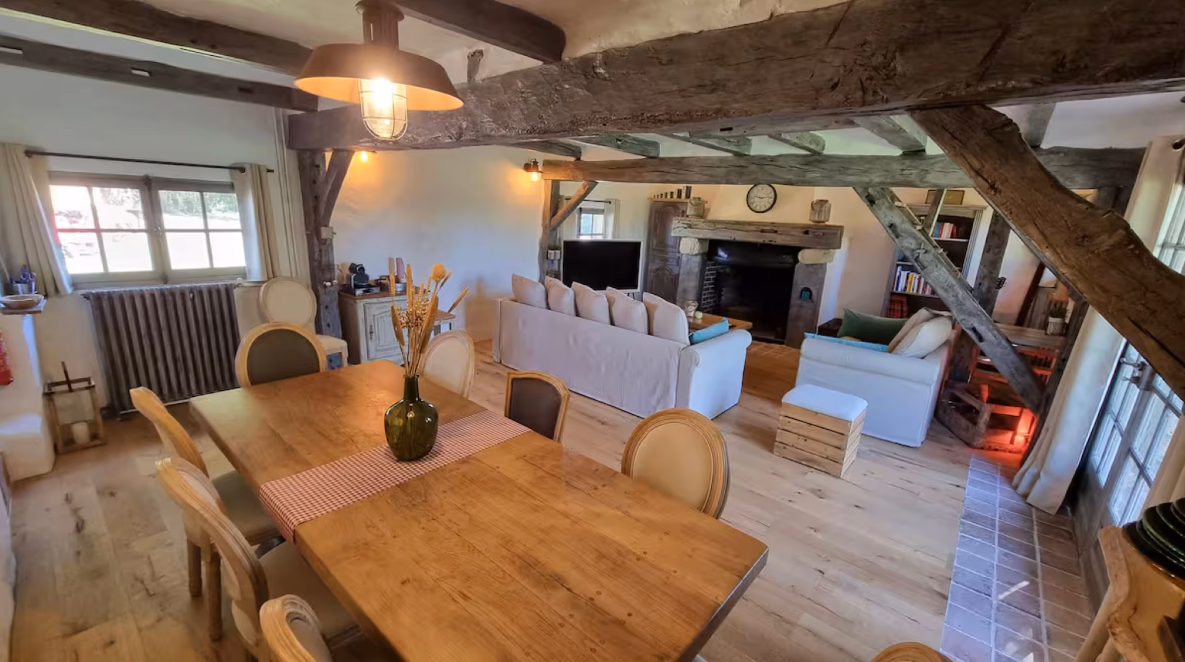 Salon ouvert avec grosses poutres apparentes en chêne, canapés crème face à la cheminée fonctionnelle et grande table en bois au premier plan.