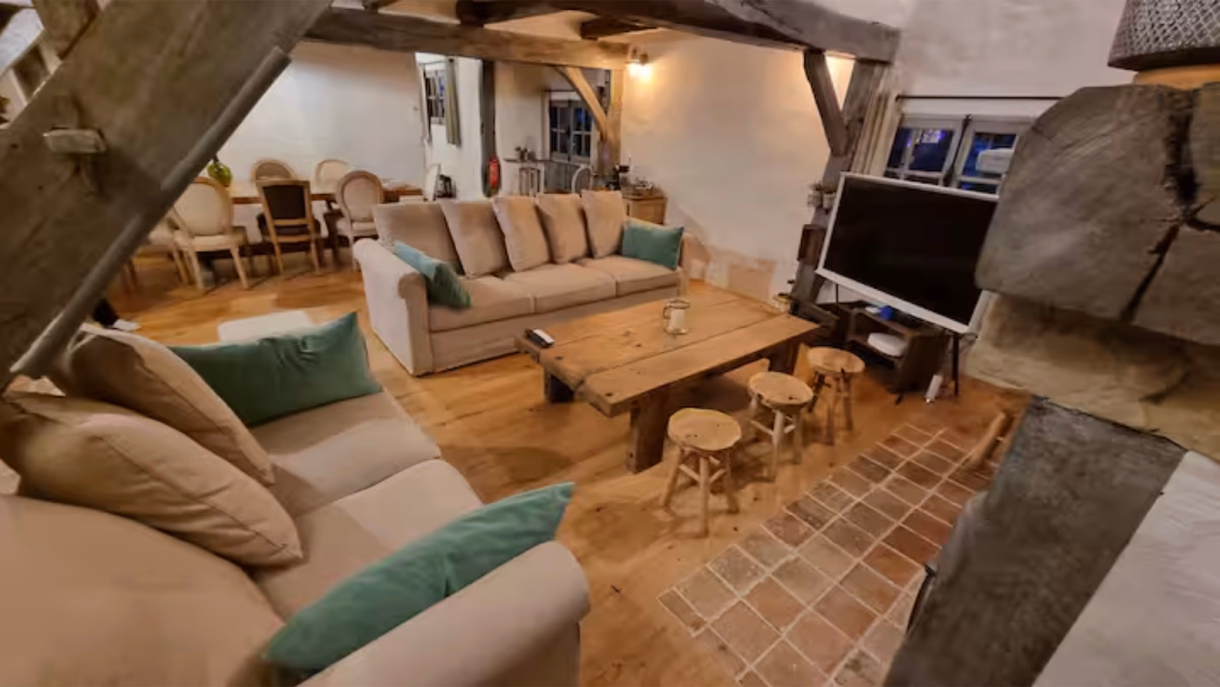 Vue large du salon : grand canapé d'angle, table basse rustique en bois brut, télévision et tabourets en bois sur sol en parquet et carreaux anciens.