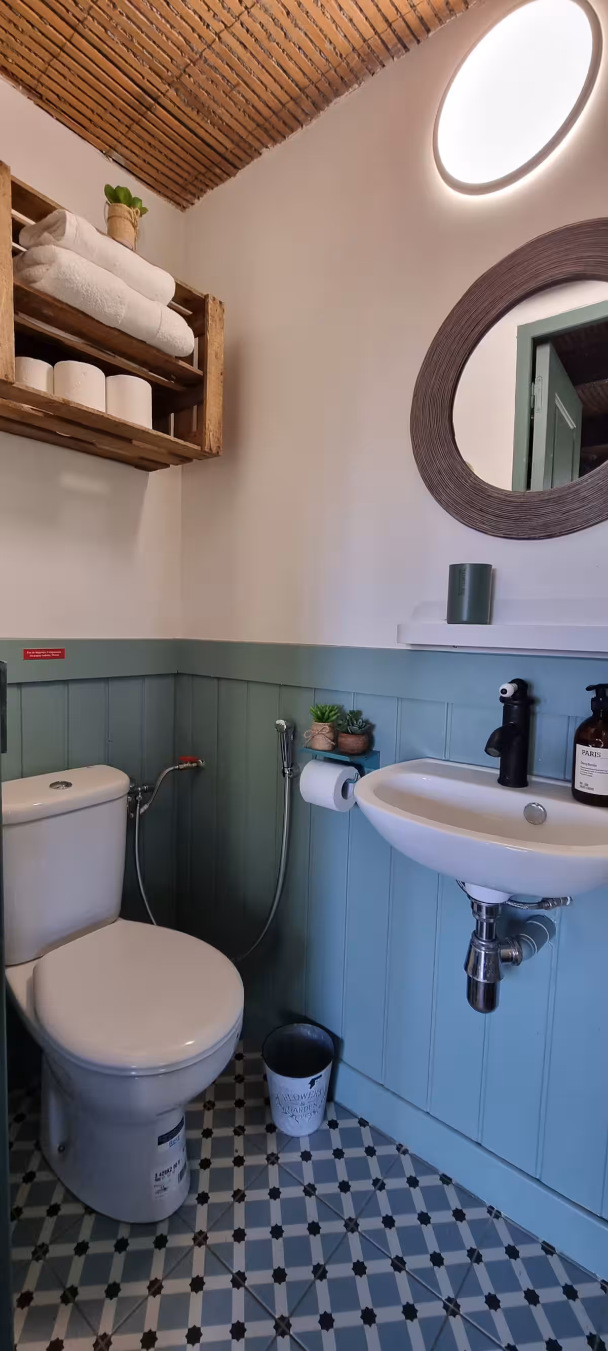 Toilettes indépendantes avec lambris turquoise, lavabo suspendu, miroir rond et plafond en bambou tressé.